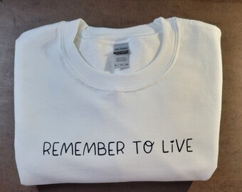 Puede incluir: Sudadera blanca con la frase "REMEMBER TO LIVE" en negro en la parte inferior. La sudadera de cuello redondo está doblada. La etiqueta de la marca Gildan es visible. Prenda informal de manga larga.