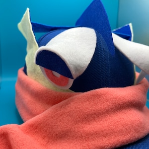 May include: A blue and white plush hat with a red and white eye and a pink scarf. The hat has a pointed top and a white brim.