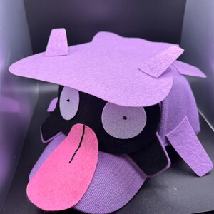 K&ouml;nnte beinhalten: Ein handgefertigter Hut aus lila Filz in Form einer Zeichentrickfigur. Der Hut hat gro&szlig;e wei&szlig;e Augen, ein schwarzes Gesicht, eine rosa Zunge und lila Akzente. Der Hut ist so gestaltet, dass er einer beliebten Figur &auml;hnelt.