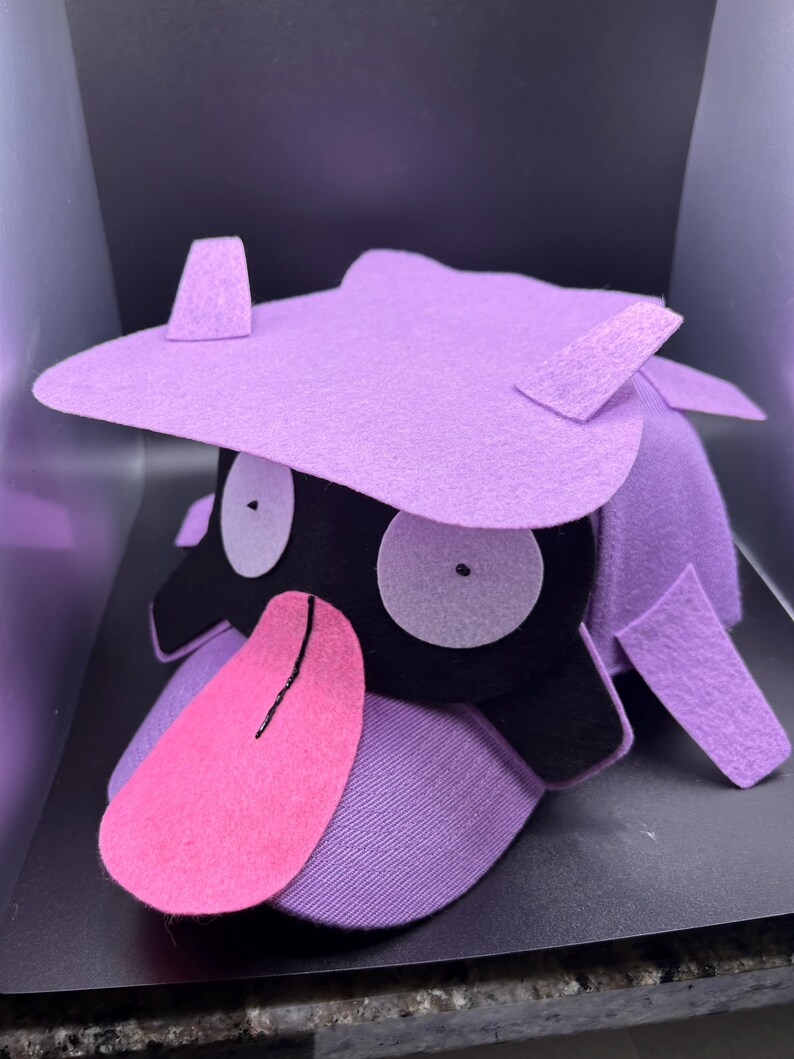 K&ouml;nnte beinhalten: Ein handgefertigter Hut aus lila Filz in Form einer Zeichentrickfigur. Der Hut hat gro&szlig;e wei&szlig;e Augen, ein schwarzes Gesicht, eine rosa Zunge und lila Akzente. Der Hut ist so gestaltet, dass er einer beliebten Figur &auml;hnelt.