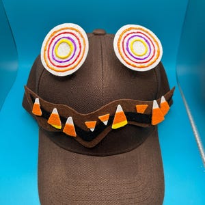 May include: A brown baseball cap with a felt monster face. The monster has a black mouth with white teeth, orange and white candy corn, and two white eyes with colourful concentric circles.