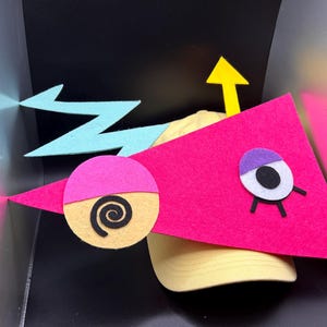 May include: A yellow baseball cap with a pink felt triangle and a light blue lightning bolt design. The pink triangle has a purple and white eye, and a yellow arrow. A pink circle with a black spiral is also attached.