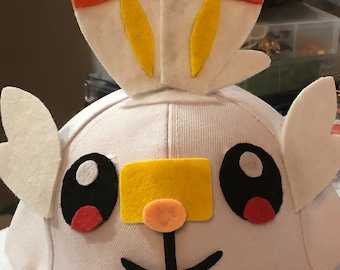 May include: White baseball cap with a felt appliqué of a cartoon character. The character has large black eyes, a yellow nose, and a black mouth. The character's ears are white with red and yellow accents.