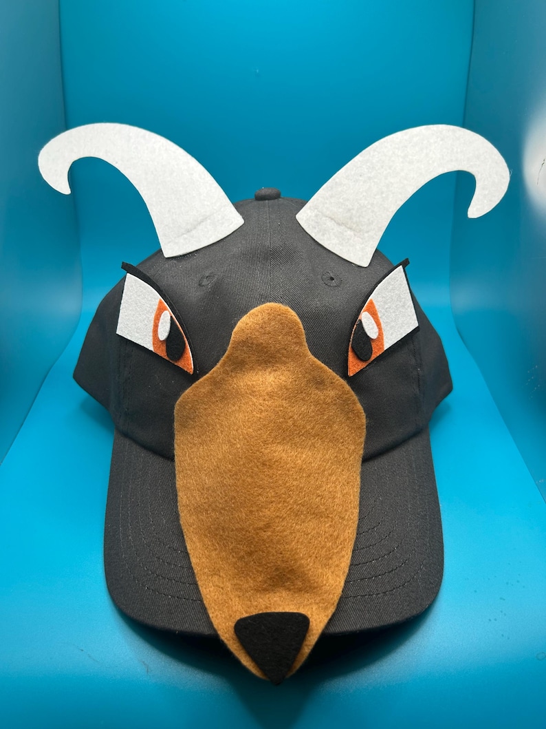 Peut inclure: Casquette de baseball noire avec des cornes en feutre blanc, des yeux orange et blancs et un museau en feutre marron. La casquette a une visi&egrave;re incurv&eacute;e et un dessus arrondi.