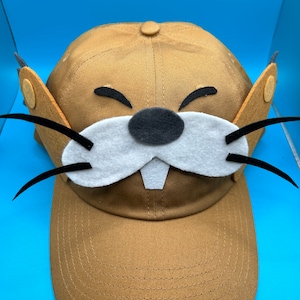 May include: A brown baseball cap with a felt applique of a cartoon animal face. The animal has a white snout, black eyes, a black nose, and black whiskers.