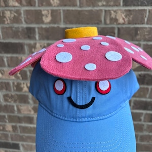 May include: A novelty blue baseball cap featuring a smiling face, red eyes, and a pink mushroom-shaped top with white spots. The mushroom top has a yellow band. A playful and distinctive accessory.