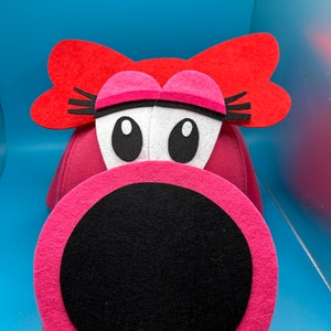 May include: Pink felt hat with a large black circle in the front, resembling a mouth. The hat has red felt ears with black eyelashes and white felt eyes.