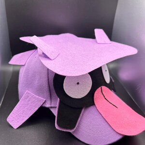 K&ouml;nnte beinhalten: Eine lila Baseballkappe mit einem Cartoon-Entenschnabel-Design. Die Kappe hat einen helllila Schirm, einen schwarzen Schnabel und eine rosa Zunge. Die Augen sind wei&szlig; mit schwarzen Pupillen. Der Hut hat ein einzigartiges, skurriles Design.