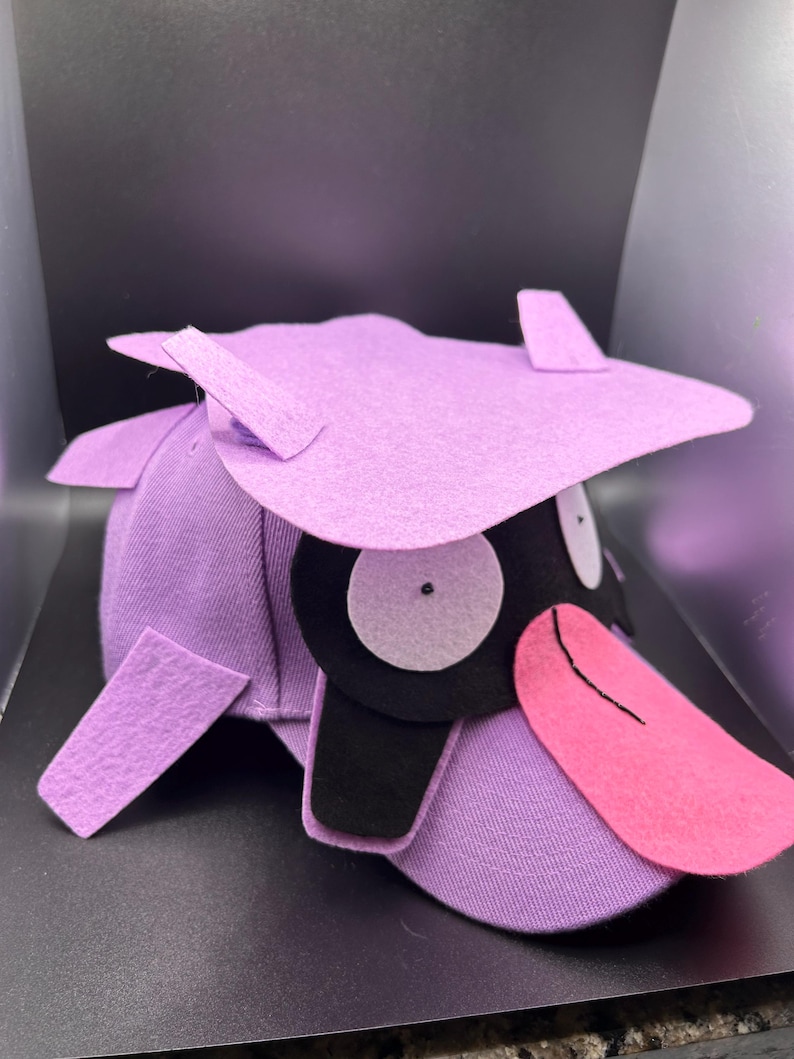 K&ouml;nnte beinhalten: Eine lila Baseballkappe mit einem Cartoon-Entenschnabel-Design. Die Kappe hat einen helllila Schirm, einen schwarzen Schnabel und eine rosa Zunge. Die Augen sind wei&szlig; mit schwarzen Pupillen. Der Hut hat ein einzigartiges, skurriles Design.