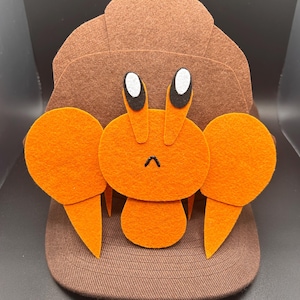May include: A brown baseball cap with a felt crab design. The crab has orange claws, eyes, and body. The cap is made of a brown fabric and has a curved brim. The crab's eyes are white with black accents.