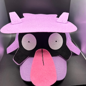 May include: A handmade plush toy in the shape of a cartoon character. The toy features a lavender head and body, black face, large white eyes, and a pink tongue. The character has a hat and side flaps.