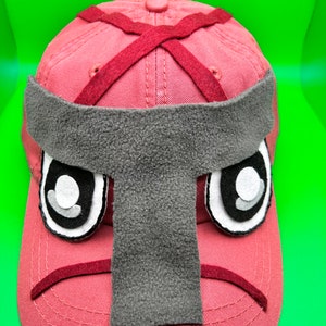 May include: A pink baseball cap with a gray felt face mask. The mask has two black eyes with white pupils and a frown.