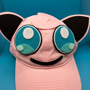May include: Pink baseball cap with a pink and black felt applique of a Pokemon character with large blue eyes.