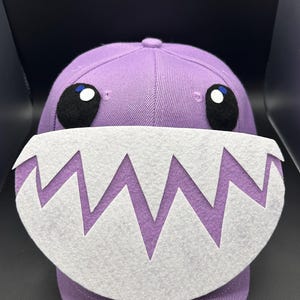 May include: A purple baseball cap with a cartoon monster design. The cap has black eyes with white and blue accents, and a white felt mouth with jagged teeth. The hat is made of a textured fabric.
