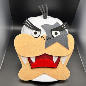 May include: A white baseball cap with a cartoon character design. The cap features a tan face, red mouth, white teeth, and black and grey accents. The design resembles a video game character.