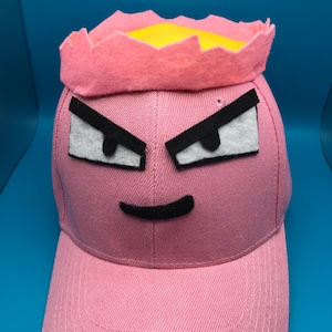May include: Pink baseball cap with a yellow crown and a black and white angry face.