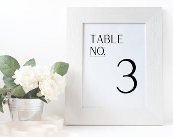 May include: White framed table number sign with black text that reads "TABLE NO. 3". The sign is sitting on a white surface with a bouquet of white roses in a silver bucket.