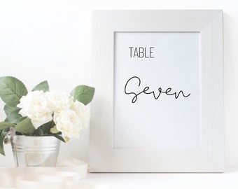 May include: White framed table number sign with the text "TABLE Seven" in black script font.