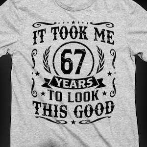 Puede incluir: Camiseta gris claro con un gráfico negro que dice: "IT TOOK ME 67 YEARS TO LOOK THIS GOOD". El diseño incluye el número 67 dentro de un círculo, con elementos decorativos.