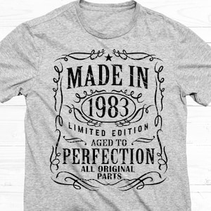 Op de afbeelding: Een grijs t-shirt met een zwart grafisch ontwerp. Het ontwerp bevat de tekst "MADE IN 1983" in een decoratief kader, met "LIMITED EDITION" en "AGED TO PERFECTION" eronder. Het is een casual kledingstuk.