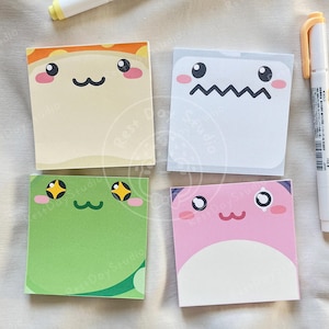 May include: Four square sticky notes with cartoon faces in beige, white, green, and pink. Two white pens with yellow and orange accents are also in the image. The sticky notes feature cute, expressive faces.