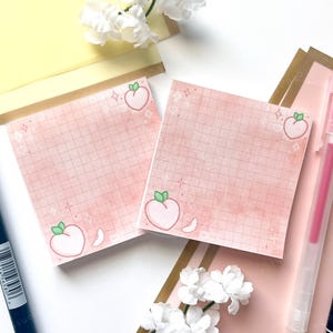Pode incluir: Duas notas adesivas quadradas rosa com uma ilustração de pêssego e um padrão de grade. As notas são cercadas por canetas rosa e brancas e flores brancas. O fundo é uma mistura de rosa e amarelo.