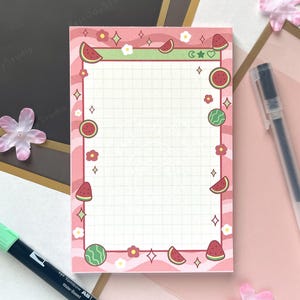 May include: A pink and white grid-lined notepad with a watermelon theme. The notepad is decorated with watermelon slices, flowers, and green accents. Two pens, one black and one pink, and a green marker are also visible.