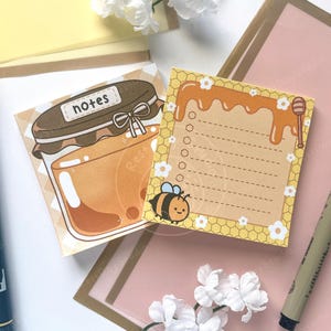 Könnte beinhalten: Zwei illustrierte Haftnotizen mit Honig-Motiv. Eines zeigt ein Honigglas mit dem Wort "Notizen". Das andere hat eine Biene und Honig, der von oben tropft. Ein schwarzer Stift und ein gelber Marker sind ebenfalls im Bild.