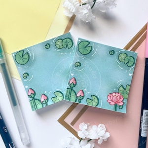 May include: Two square notepads with a watercolor illustration of a pond with lily pads and lotus flowers. The pads are light blue with green and pink accents. Two pens are on either side of the notepads.