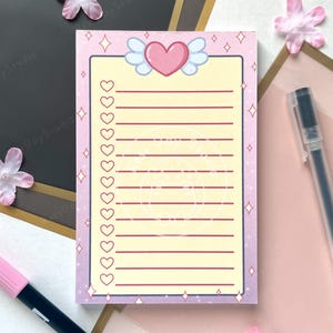 May include: A pastel pink and purple notepad featuring a heart and wing design. The notepad has a list of lines with heart-shaped bullet points. Pens and decorative flowers are in the background.