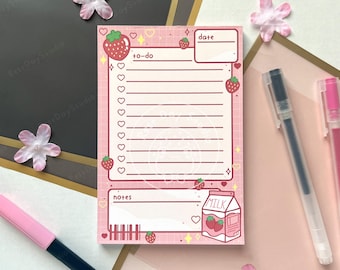 May include: A pink to-do list notepad with a strawberry theme. The notepad features a grid pattern, strawberry illustrations, and the words "to-do", "date", and "notes". A milk carton with strawberries is also shown. Pens and decorative flowers are in the background.
