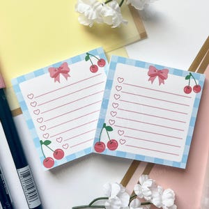 May include: Two square notepads with a blue gingham border, pink bows, and red cherries. Each notepad has lined pages and a row of hearts. Pink and blue markers and a red pen are also visible.
