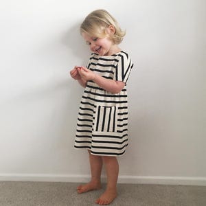 May include: A striped dress in black and cream, worn by a child. The dress features horizontal stripes on the body and vertical stripes on the pocket. The child is smiling and barefoot, against a white wall.