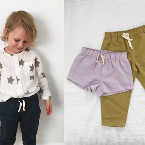 May include: A white long-sleeved shirt with a brown star pattern, a pair of dark navy pants with a white drawstring, a pair of olive green pants with a white drawstring, and a pair of light purple shorts with a white drawstring waistband.