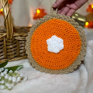 May include: A crocheted pumpkin pie pot holder with a white star in the center. The pot holder is orange and brown and has a loop for hanging.
