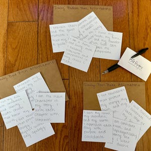 May include: A collection of handwritten affirmation cards on a wooden surface. The cards are grouped with titles like "Daily Badass Mom Affirmations" and "Daily Boss Babe Affirmations." A pen and a business card are also present.