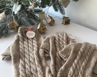 May include: Two beige knitted dog sweaters with cable knit pattern and short sleeves. The sweaters have a wooden tag that says "Handmade with love".