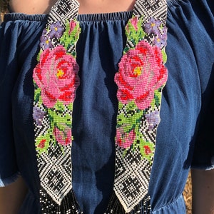 Puede incluir: Un collar largo de cuentas con un diseño floral. El collar está hecho de cuentas blancas y negras y presenta flores rosas, rojas y verdes. El collar tiene una franja de cuentas negras.