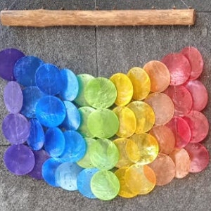 May include: A rainbow-colored wind chime made of iridescent seashells. The chime is hanging from a wooden dowel and is made up of multiple rows of shells in shades of purple, blue, green, yellow, and red.