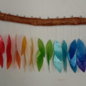 May include: A colorful wind chime with a natural wood branch and iridescent leaf-shaped pendants. The pendants are arranged in a rainbow spectrum, with colors ranging from red to violet. The chime hangs against a white wall.