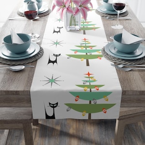 May include: White table runner with a mid-century modern design featuring stylized green Christmas trees, black cat silhouettes, and starbursts. The trees are adorned with colorful ornaments. The runner is set on a wooden table with place settings.