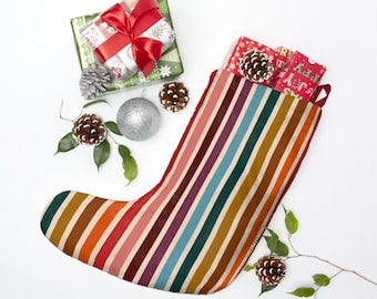 May include: A Christmas stocking with vertical stripes in various colors, including orange, red, and teal. The stocking is filled with wrapped gifts and pine cones. Additional gifts, ornaments, and pine cones are arranged around the stocking on a white surface.