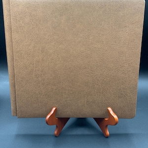 May include: A brown leather-bound photo album, displayed on a wooden stand. The album has a textured surface and a rectangular shape. The stand is made of wood and has a curved design. The album is closed, and the pages are visible at the top.