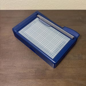 May include: A dark blue paper trimmer with a gray grid cutting surface. The trimmer has a silver blade guide and a dark blue base. The grid is marked with measurements. The trimmer is on a brown wooden surface.