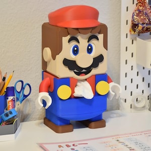 May include: A 3D printed tissue box cover shaped like a Super Mario character. The cover is brown, red, blue, and yellow. The character has a mustache and a red hat.