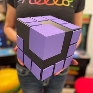 May include: A large, cubic, three-dimensional puzzle toy featuring a matte black base structure with raised, square, light purple panels arranged in a grid pattern on each visible face, mimicking the appearance of a classic mechanical puzzle.