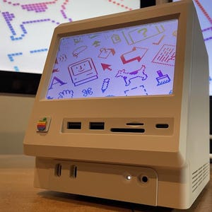 Op de afbeelding: Drie beige computerschermen in retrostijl met kleurrijke krabbels. Elke monitor heeft een scherm, een sleuf voor een schijf en een aan/uit-knop. De tekst "Cover 1", "Cover 2" en "Cover 3" staat onder elke monitor.