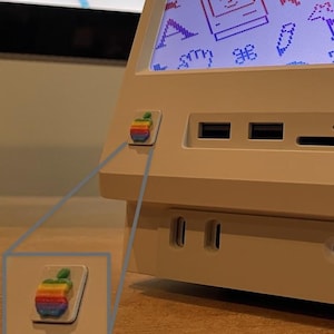 Könnte beinhalten: Ein Computer im Retro-Stil mit einem Bildschirm, der farbenfrohe Symbole und eine Hunde-Grafik anzeigt. Das cremefarbene Gerät hat ein Regenbogen-Apple-Logo und verschiedene Anschlüsse. Der Bildschirm zeigt eine Retro-Computer-Oberfläche.