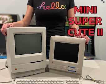 May include: Two vintage Macintosh computers, a keyboard, and a mouse are displayed. The computers are off-white with black screens. The keyboard and mouse are also off-white. The text "MINI SUPER CUTE II" is visible in the background.