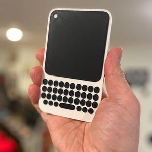 May include: A white and black handheld device with a keyboard and screen. The device has a rectangular shape with rounded corners. The screen is black, and the keyboard has black circular keys. The device is held in a person's hand.